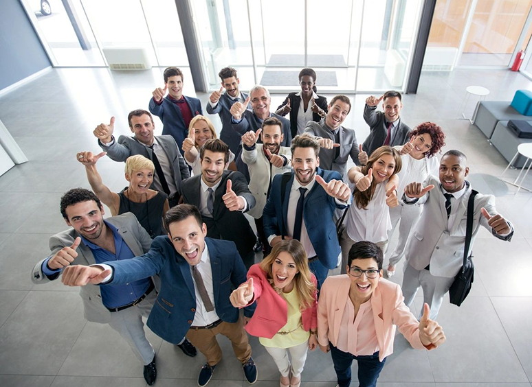 A diverse group of professionals smiling and giving thumbs up in a bright office.
